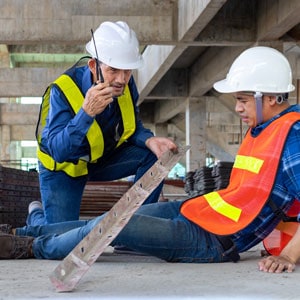 Two men in safety vests and hard hats are on the ground, discussing compensation for construction injuries. - Escamilla Law Firm, PLLC